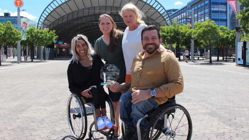 Two people in a wheelchair and two women standing smile with an award