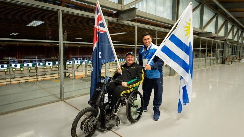 A man in a wheelchair holding the Australian flag posing next to a man standing and holding the flag of Uruguay