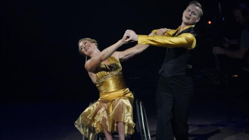 A female wheelchair dancer holding hands with her male standing dance partner on a dancefloor
