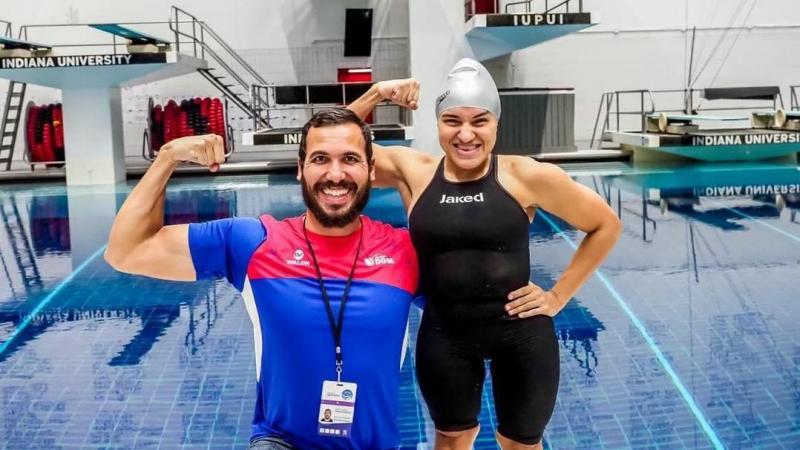 A short stature woman in swimming outfit with her arm around a knelled man with a swimming pool in the background