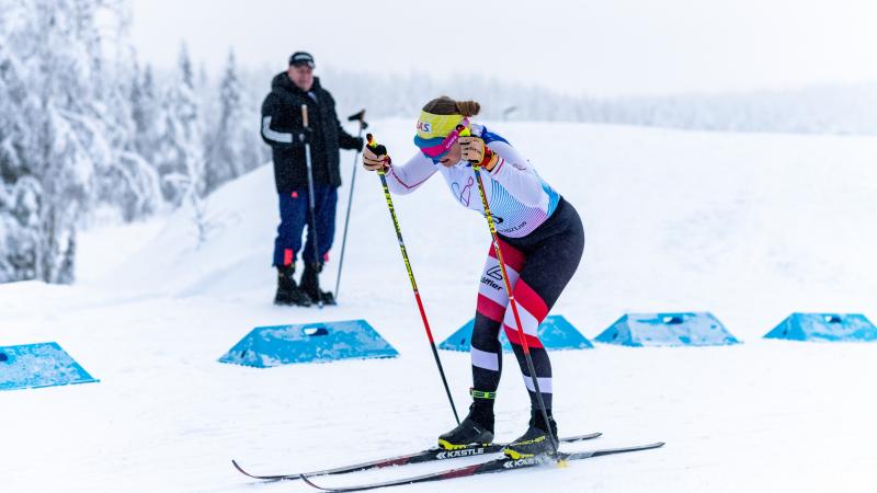 A female cross-country skier competing in the snow observed by a man in the background