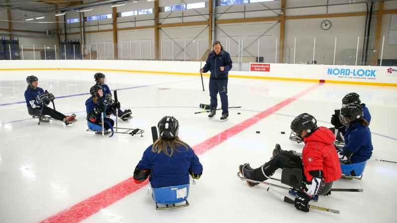 A woman on an ice rink talking to a group of seven people on Para ice hockey sledges 