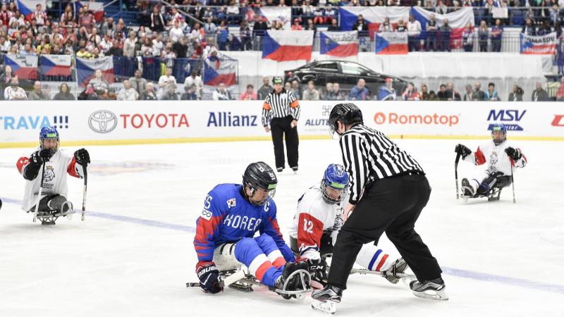 A referee throwing the puck in front of two players to start a Para ice hockey game in a crowded arena