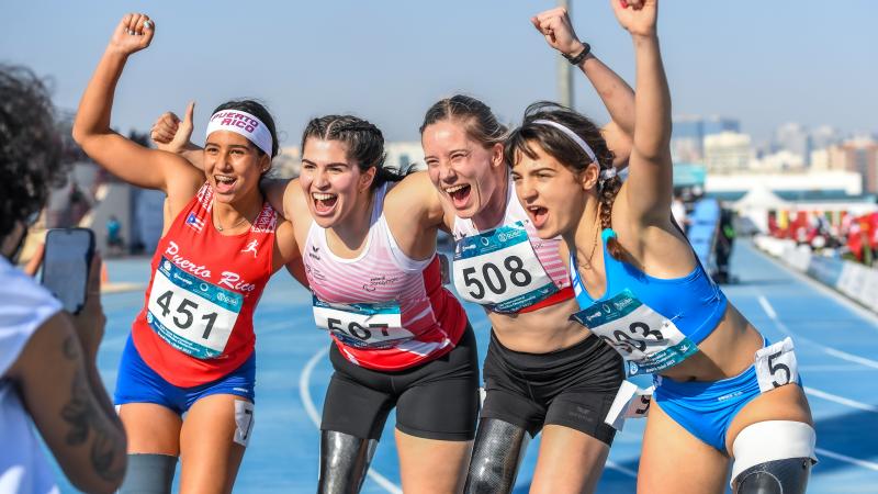 Four women with prosthetic legs hugging each other and smiling to a person taking a picture with a mobile phone