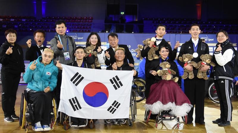 A group of 12 South Koreans, four of them in wheelchairs, posing for a group photo after the Bonn 2019 World Para Dance Sport Championships