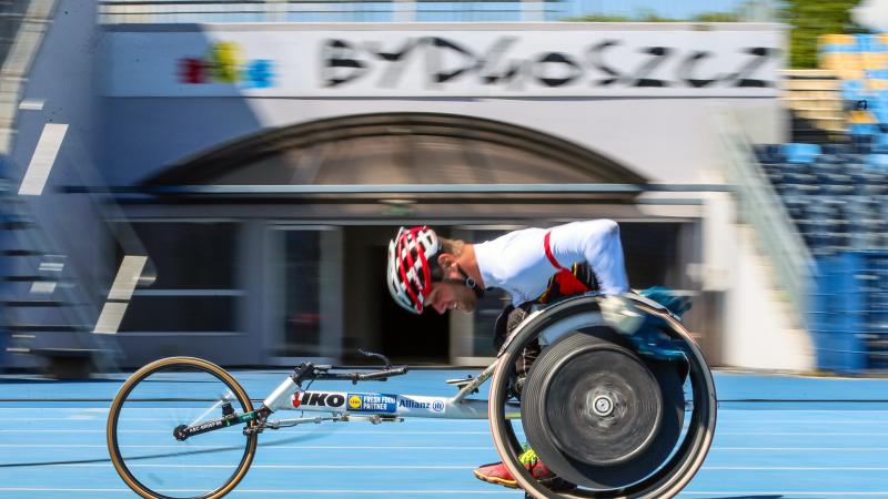 A man in a racing wheelchair on a blue athletics track passing in front of a gate