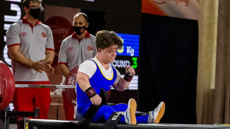 A female powerlifter celebrating seated on the bench press with two men standing behind her