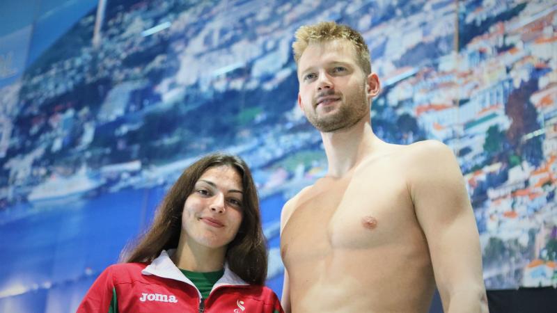 A short woman with a Portugal jumper and a tall shirtless man side by side in front of a panel in a swimming pool