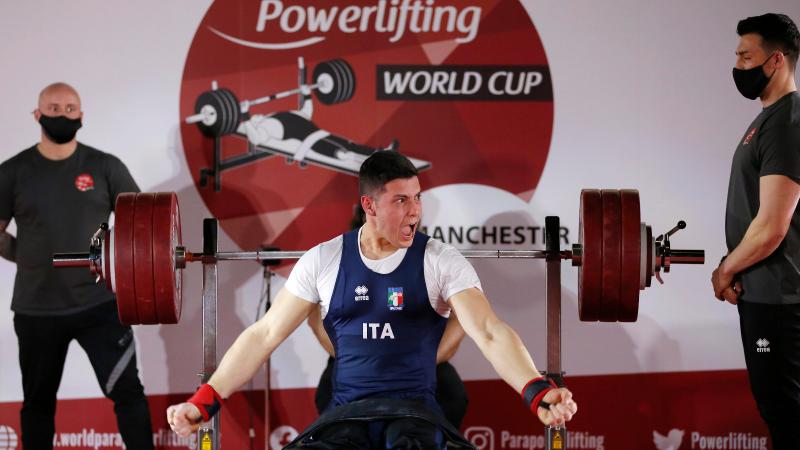 A men without legs on a bench press celebrating observed by two other men standing during a Para powerlifting competition