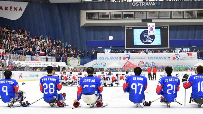 Two Para ice hockey teams on ice looking at a giant screen in a packed arena