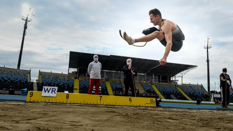 A men with a prosthetic leg jumping in a long jump event observed by three man
