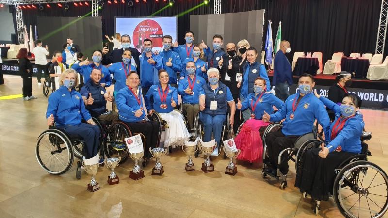 A group of nine people in wheelchairs with 12 people standing behind them posing for a picture with trophies in front of them