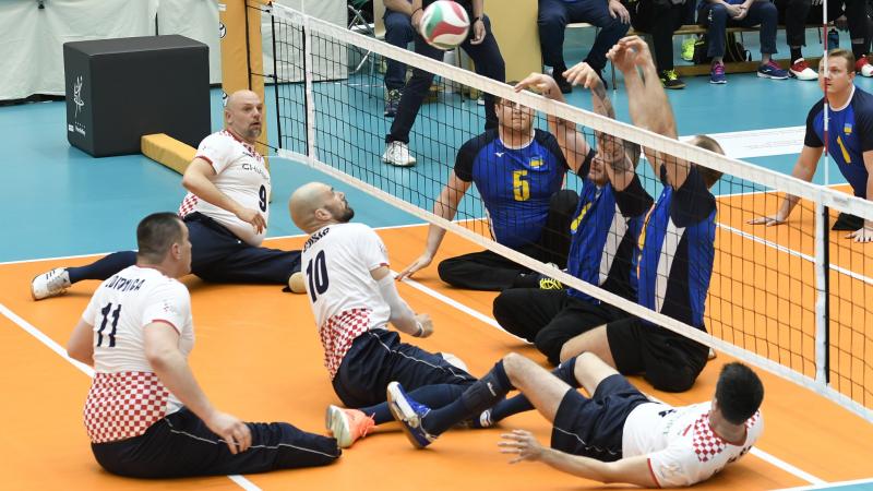 Croatian and Ukrainian sitting volleyball players separated by the net trying to reach the ball
