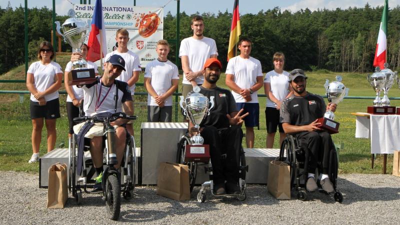 Three men in wheelchairs holding trophies with seven persons standing behind them