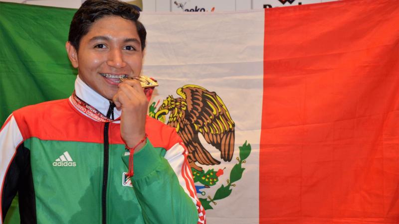 Male Mexican taekwondo athlete smiles biting medal in front of Mexican flag
