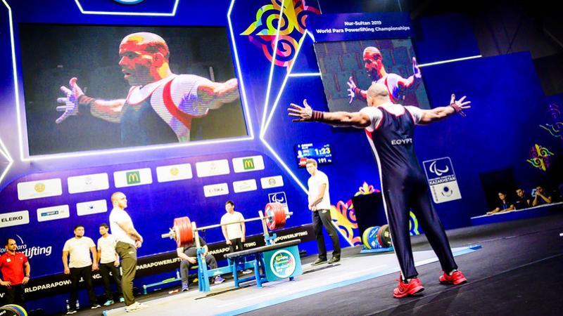 A man standing in front of a bench press on a stage observed by six men standing and three persons behind a table