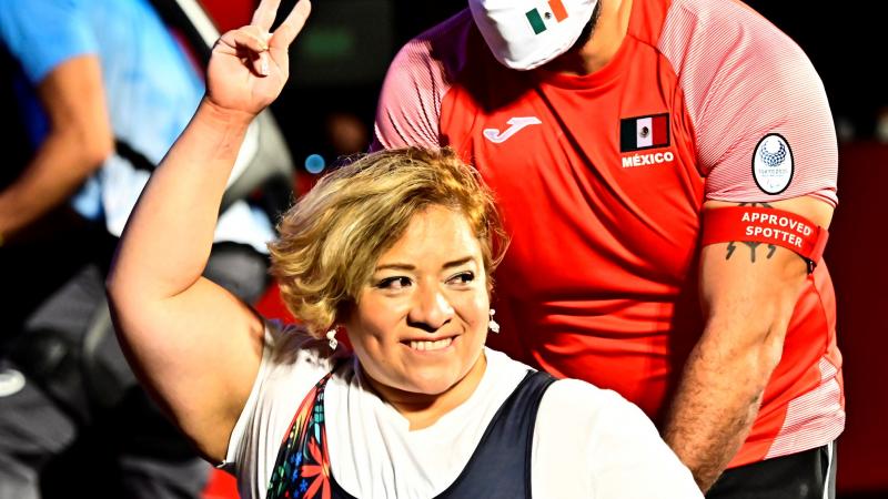 A woman smiling and raising her arm in a wheelchair chair being pushed by a man