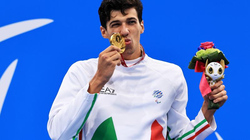 A man kisses the gold medal with a smile on his face. He is holding the miniature mascot in the other hand