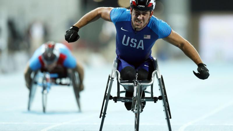 A man in a racing wheelchair celebrating with another competitor in the background