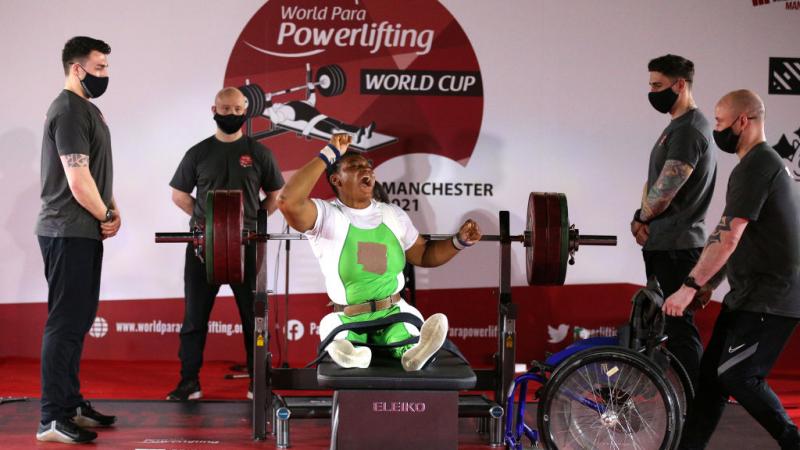 A woman celebrating on a bench press during a Para powerlifting event with four male loaders standing near her