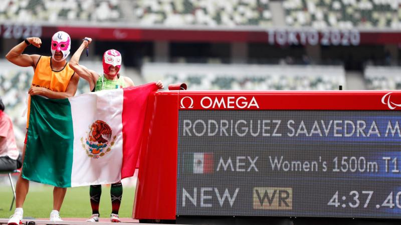 A man and a woman with Lucha Libre masks in an athletics track with the Mexican flag