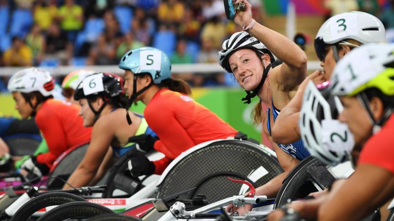 A woman in a racing wheelchair waving to the camera surrounded by other six female wheelchair racers