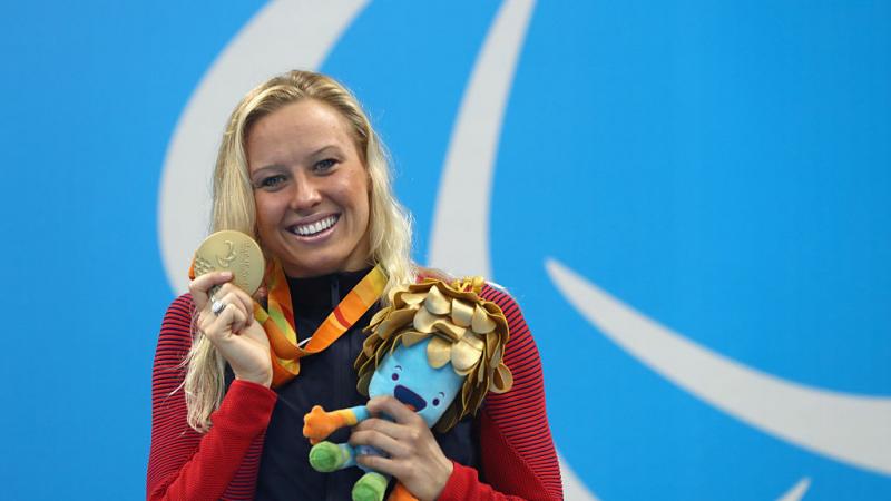 A woman posing for a photo with a smile on her face while holding a gold medal in her right hand and a mascot in her left hand