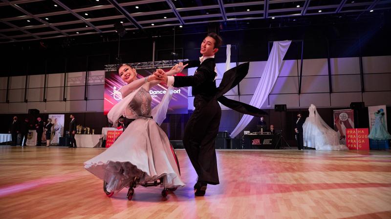 A female wheelchair dancer and her standing male partner in a Para dance sport competition observed by seven people standing in the background