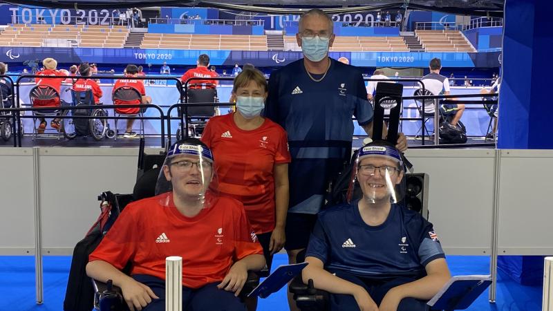 Linda stands behind Scott in his wheelchair, both wearing red, while Gary stands behind Jamie in his wheelchair, both in blue