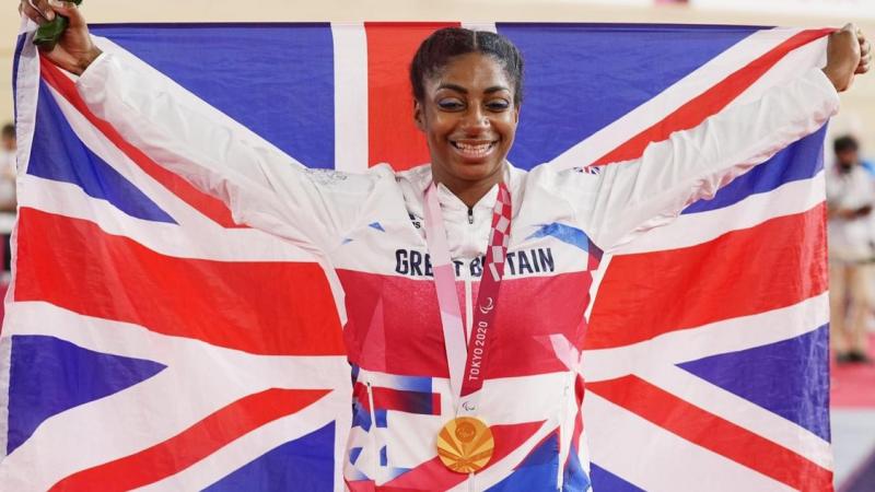 Kadeena Cox smiles with the gold medal hanging around her neck while holding the British flag