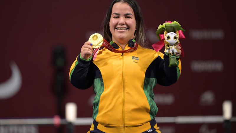 A woman posing with the gold medal in her right hand and a mascot in her left hand. She is having a big smile on her face