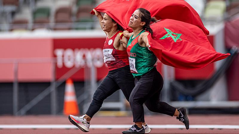 Two short stature women running on an athletics track holding the flags of Tunisia and Morocco