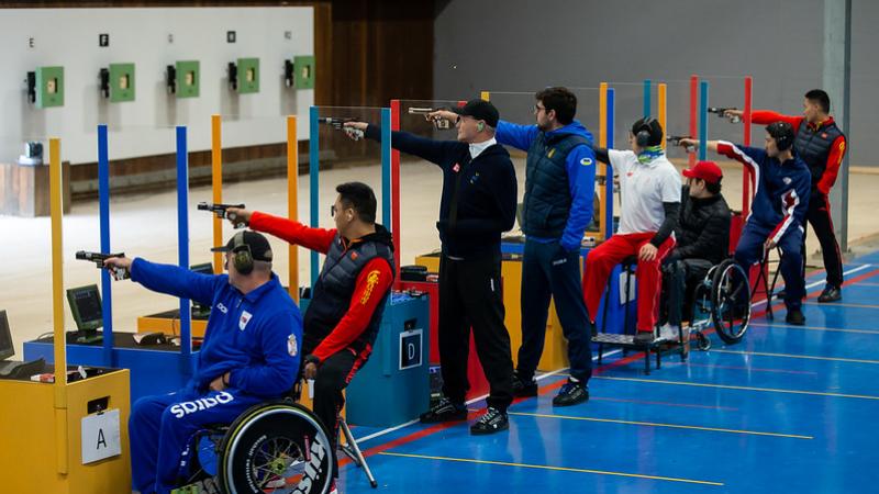 A group of eight men competing in a pistol shooting event in a shooting range