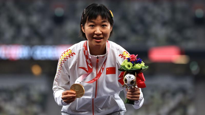 Xiaoyan Wen crouches on the podium displaying gold medal and mascot