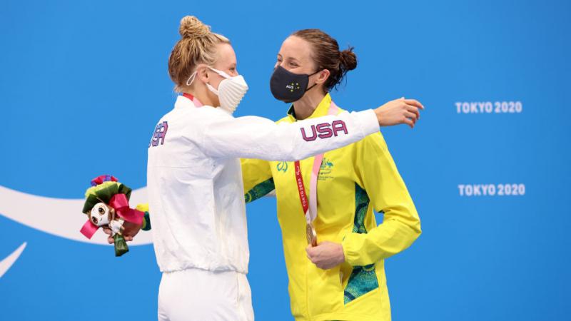Two women hugging in front of a backdrop showing the Paralympic Agitos in white