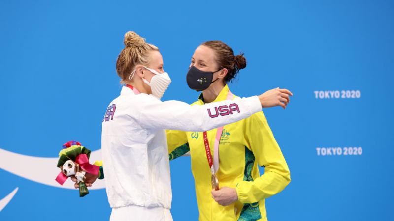 Two women hugging in front of a backdrop showing the Paralympic Agitos in white