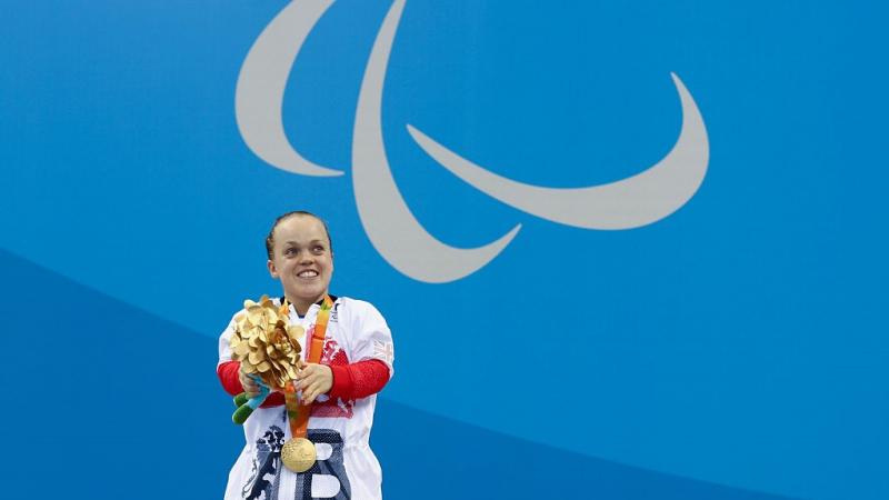 A short stature woman with her gold medal in front of a wall with the Paralympic Agitos