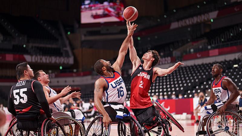 Two wheelchair basketball male athletes reach for the ball in the air