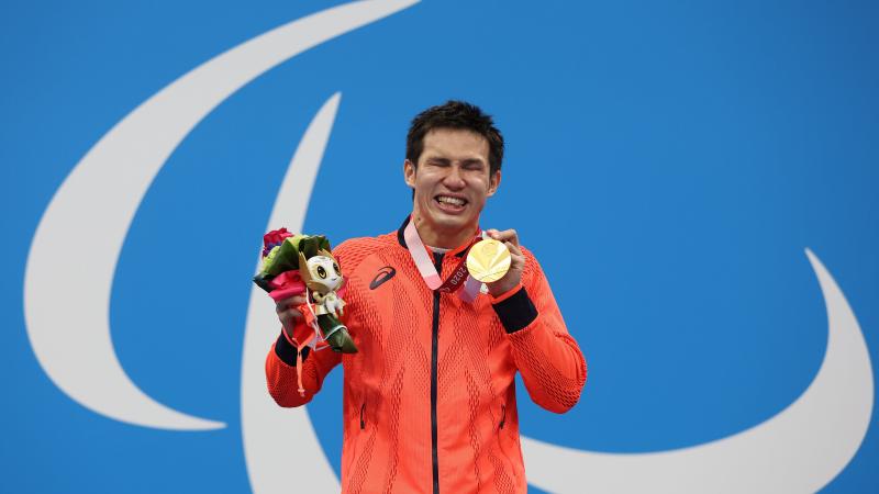 A vision-impaired athlete posing with a gold medal in left hand a mascot in his right hand