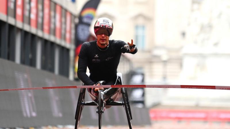 A man crosses the finish line in a wheelchair holding his left hand up with two fingers up signalizing victory