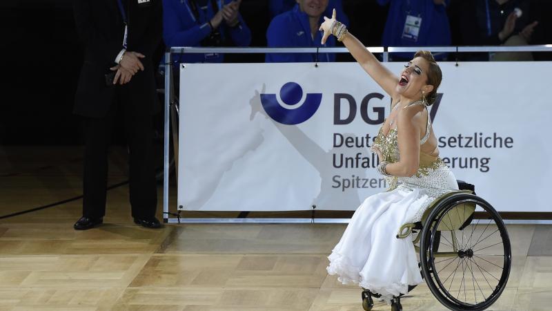 A woman in a white and gold dress dancing in a wheelchair with right arm raised