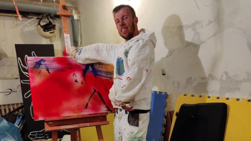 A man posing for a photo in front of his painting in a messy basement