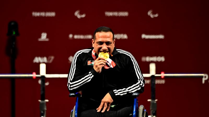 A man in a wheelchair biting the gold medal with a smile on his face