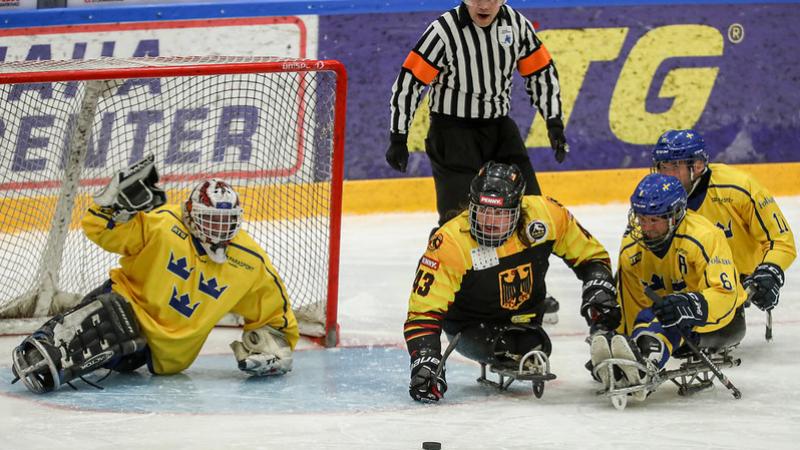 A German Para ice hockey player on ice competing against three Swedish players observed by a referee