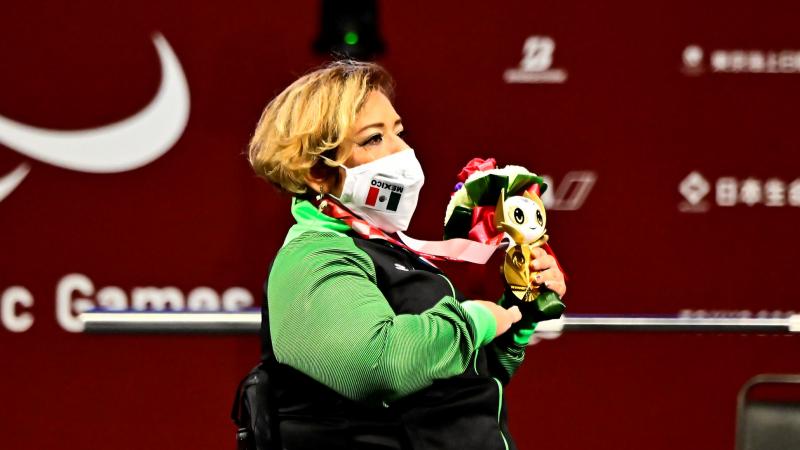 A woman in a wheelchair listening to the national anthem and holding a gold medal and a mascot in her left hand and holding her left hand high.