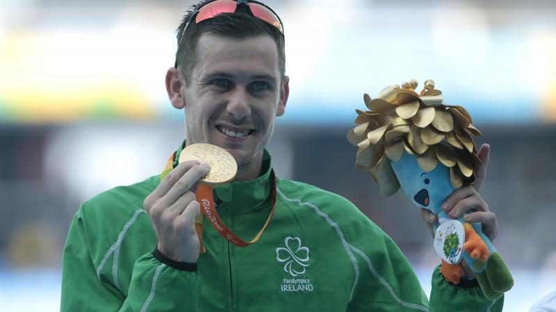 A man with the uniform of Ireland showing his gold medal and a mascot