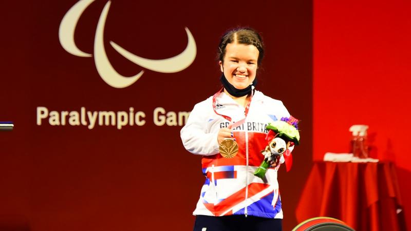 A woman holding a bronze medal in her right hand and posing for a photo on the podium.