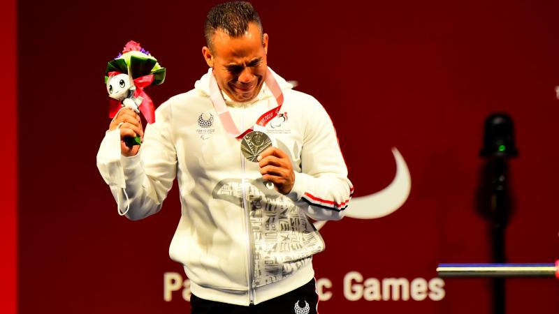 A man crying and holding a silver medal in his left hand with a mascot in his right hand.