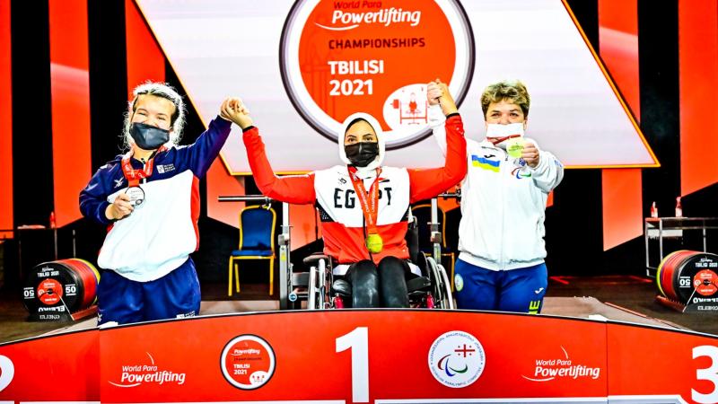 A woman in a wheelchair racing holding the arms of the short-stature women on a podium