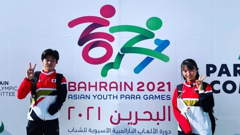 Two young Japanese Para athletes waving in front of a poster of the Asian Youth Para Games in Bahrain